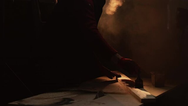 Woodwork craft. DIY joinery. Carpentry production. Unrecognizable workman painting wood with paintbrush at dark cold workshop with vapor.