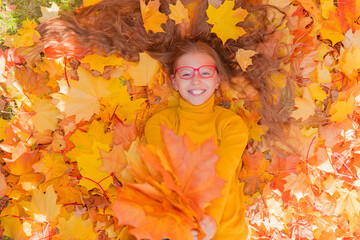 a teenage girl in red glasses lies in yellow autumn foliage and smiles