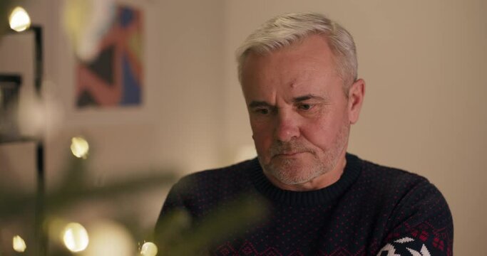 Close-up of middle-aged man with gray hair standing and looking at Christmas tree. A man dressed in sweater. He is sad, unhappy and does not want to celebrate Christmas, because he broke up with wife.