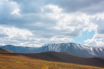 Motley autumn landscape with hills on high plateau and sunlit snowy mountain range under dramatic cloudy sky. Vivid autumn colors in mountains. Sunlight and shadows of clouds in changeable weather.