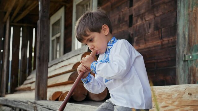 Little boy playing on woodwind wooden flute - ukrainian sopilka. Folk music concept. Musical instrument. Kid in traditional embroidered shirt - Vyshyvanka. Child sitting on porch of old wooden house