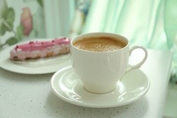 Cup of delicious aromatic coffee and eclair on white table indoors, closeup