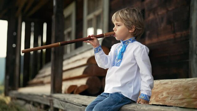 Little boy playing on woodwind wooden flute - ukrainian sopilka. Folk music concept. Musical instrument. Kid in traditional embroidered shirt - Vyshyvanka. Child sitting on porch of old wooden house