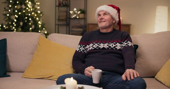 A middle-aged man sits on sofa, holding cup of tea, looking around and admiring Christmas decorations. The man takes sip, looks at the camera and smiles. He is wearing warm sweater and Santa hat.