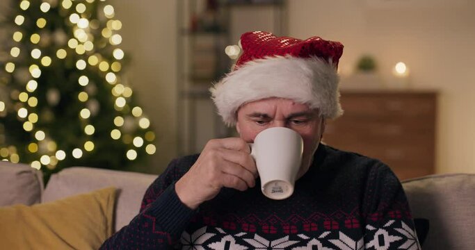 Close-up of middle-aged man sitting on sofa and wearing Santa hat. The man took cup of tea and took sip. Then he looked at the camera. He is upset because he was left alone for Christmas.