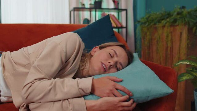 Tired caucasian adult girl lying down in bed taking a rest at home. Carefree young woman napping, falling asleep on comfortable orange sofa with pillows. Closed her eyes enjoy daytime nap alone