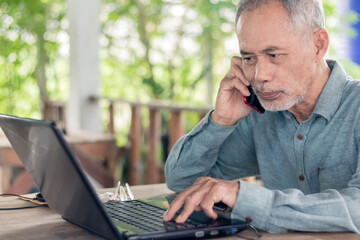 Close-up of cheerful elderly man sitting at a table talking on his mobile phone in front of his laptop.