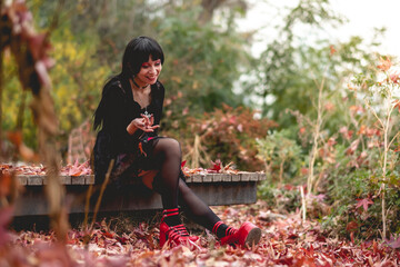 Young and skinny hispanic goth girl with black dress and red platforms shoes seated in a wood...
