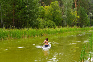 A woman drives on the Sup Board through a narrow canal surrounded by dense grass. Active weekend vacations wild nature outdoor. A woman is sitting with her legs stretched out.