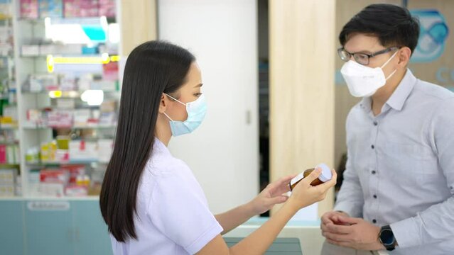 Asian woman pharmacist wear surgical face mask medication recommendation about medicine, drugs and supplements to male patient customer in drugstore. Medical pharmacy and healthcare providers concept.