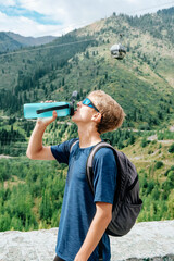 Young traveller with backpack stands on the backdrop of a stunning view with green mountains and drinks clear water from a blue bottle. High quality photo