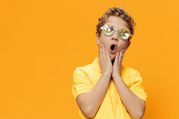  school age boy is standing in a yellow polo in funny, shiny party glasses with a very surprised expression on his face and holding his head with his hands. Horizontal studio photo with empty space