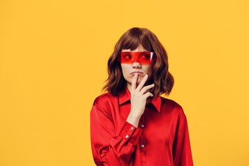 a sweet, thoughtful woman stands in a stylish red shirt and red glasses on a yellow background, thinking and holding her hand near her face. Horizontal Studio Photography