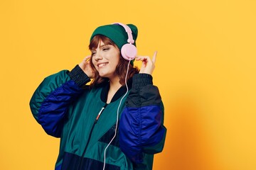 a happy, joyful woman in a green jacket and hat enjoys music standing in pink headphones on a yellow background