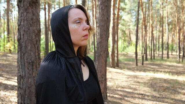 An adult woman in a black cloak with a hood stands in a pine forest. Slow motion portrait. Halloween woman portrait