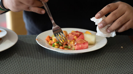 Cropped image of a young casual man eating fresh vegetables and fruits for breakfast. Right hand is holding a fork and left hand is holding a tissue.