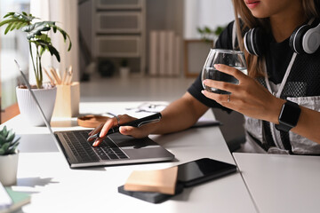Cropped view of young female freelancer holding glass of water and typing on laptop, sitting in bright home office