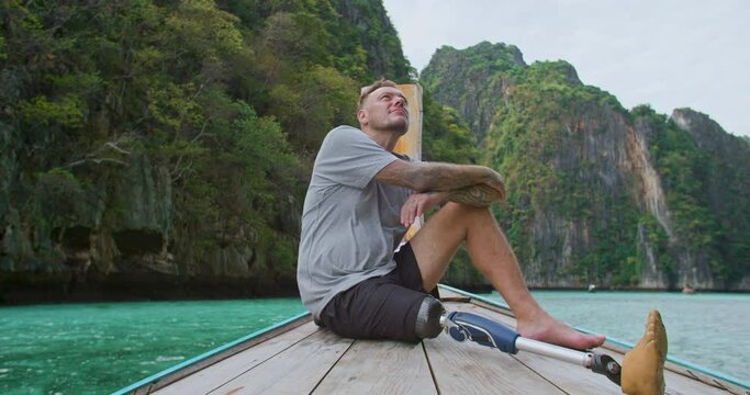 Young caucasian male with artificial bionic legs prosthesis sitting on the front of long tail boat seeing an amazing beautiful seascape during summer vacation travel in Phi Phi Islands, Thailand.