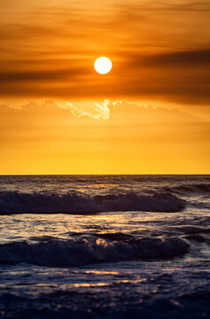 Portrait view of the orange sun rises on the ocean while the waves roll in.