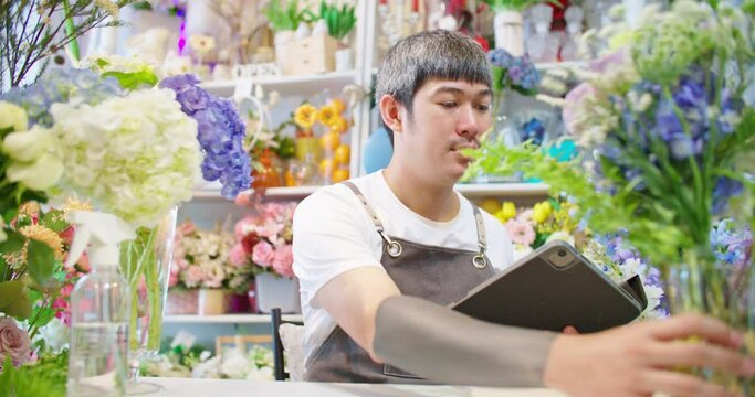 Asian male florist shop owner in apron working in store. Tattooed man hipster guy seller taking picture for customer and sending photo to client. E-commerce, SME entrepreneur, Small business online.
