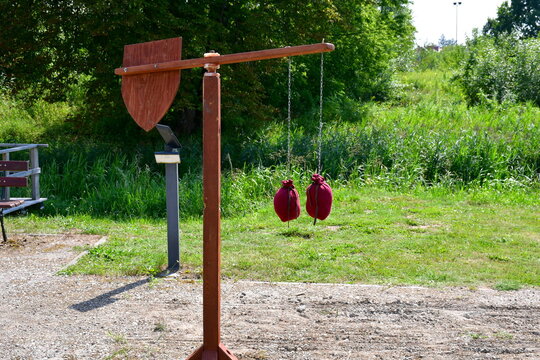 A close up on a wooden stand with a small shield or targe attached to its one arm and two pouches filled with sand attached to another seen next to a vast field or meadow on a sunny summer day