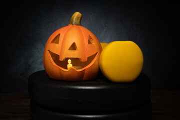 Small ceramic Halloween pumpkin with dumbbell on a barbell weight plates. Healthy fitness lifestyle autumn fall composition. Decorative Jack-o'-lantern spooky head. Gym workout and training concept.