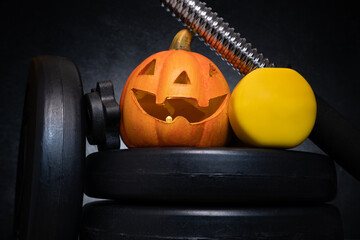 Small ceramic Halloween pumpkin with dumbbell on a barbell weight plates. Healthy fitness lifestyle autumn fall composition. Decorative Jack-o'-lantern spooky head. Gym workout and training concept.