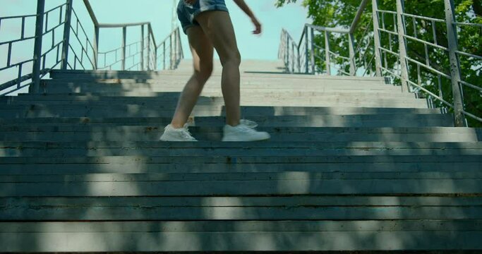 Happy young woman dancing on the stairs and enjoying life, view of the blue sky with green treetops. Carefree, youth culture and city life concept