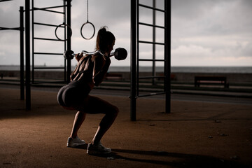 young athletic woman standing on bent knees and holding barbell on her shoulders. - obrazy, fototapety, plakaty