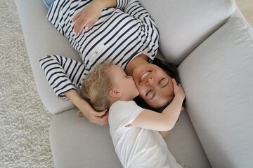 Kid girl daughter kiss smiling mom on cheek, hugging, lying on couch at home. Happy motherhood