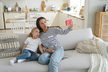 Little daughter, mom show tongues, take selfie photo by smartphone, sitting on sofa together at home
