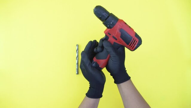 close up footage hand wearing black gloves inserting battery and drill bit to red cordless drill isolated yellow background