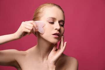 Beautiful young woman doing facial massage with gua sha tool on pink background