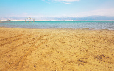 Fototapeta na wymiar Calm day at Ein Bokek Dead Sea beach, blue green water, sun shade shelter near, sun shines on sandy beach shore