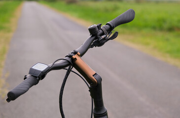 Fototapeta na wymiar Konzept Fahrradfahren allein in der Natur: Nahaufnahme eines Fahrradlenkers eines E-Bikes im Vordergrund, Fahrradweg und grüne Natur im verschwommenen Hintergrund