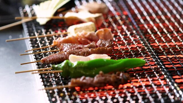 炭火の熱気で香ばしく焼き上がる串焼き。牛串や鶏肉、ししとうなどの具材を網の上で丁寧に焼き上げる調理シーン。食欲をそそるシズル感とライブ感あふれる映像で、居酒屋やバーベキューのメニュー紹介に最適な動画素材です。 | Sizzling assorted skewers being grilled on a charcoal net.