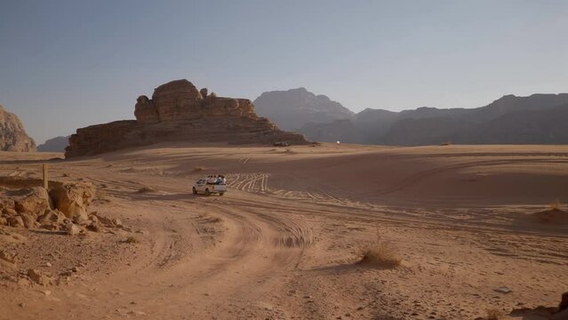 Off-road 4x4 SUV with tourists sit down on the rear body follows the tracks on the red sand of the Wadi Rum desert.