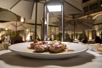 A plate of Iberian pork meat ,Iberian feather. The Iberian feather is the triangular-shaped part of the pig located at the back of the loin. Outside a restaurant, illuminated by a modern table lamp. 