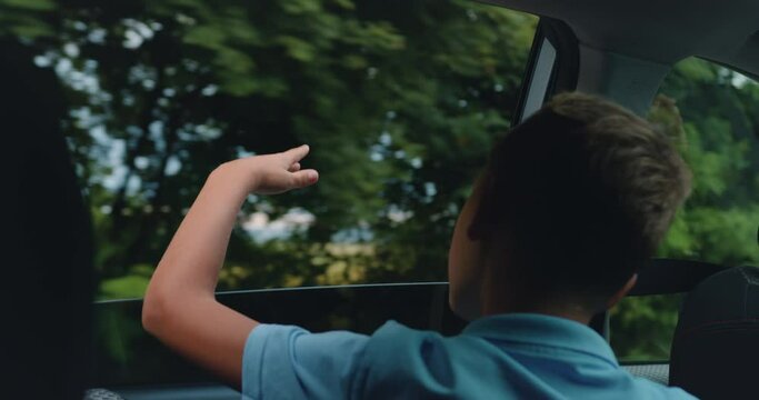 Back view. Happy child stretched out his hand from the car window. Boy waving hands gesturing out of the car window during a trip with his family.