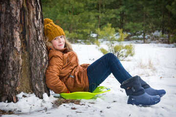 In the winter afternoon in a snowy pine forest, a girl sits on a plastic ice plate.