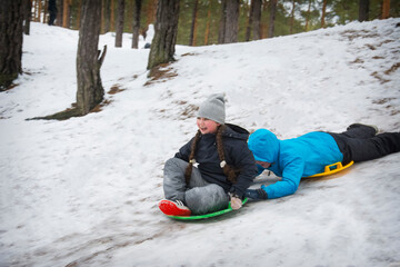 In winter, in a pine forest on a bright  day, little brother and sister ride from the mountain on plastic plates.