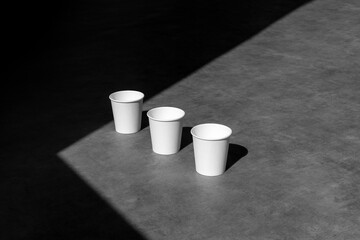 A row of blank coffee paper cups mockup, front view, on concrete background, with sharp natural sunlit shadows. Packaging mockup template with empty space to display your branding design.