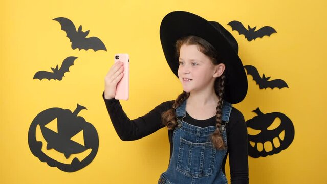 Cute little kid girl in hat get video call using mobile cell phone talk greet with hand, isolated on yellow color background in studio decorated paper black pumpkins and bats. Happy Halloween concept