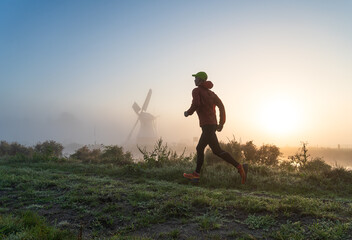 Man running in the foggy, Dutch countryside near a windmill during a tranquil sunrise. With motion...