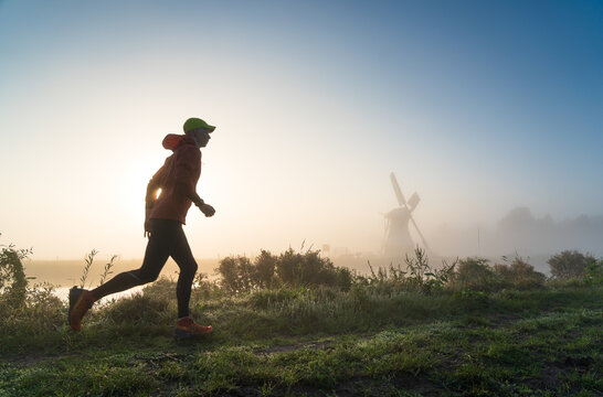 Man running in the foggy, Dutch countryside near a windmill during a tranquil sunrise. With motion blur.