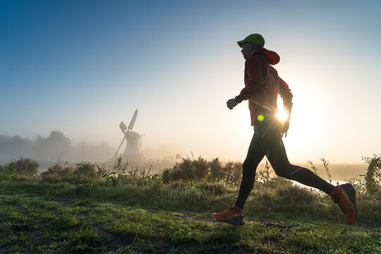 Man running in the foggy, Dutch countryside near a windmill during a tranquil sunrise. With motion blur.