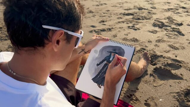 Male artist sitting on a sand beach painting chracter with watercolor and paintbrush. Game developer 2D artist making up design for videogame. Man drawing outdoors. Film grain. Soft focus. Live camera