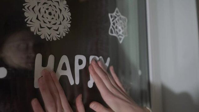 Teenage girl sticks paper word Happy on window glass. Christmas and new year holiday concept.