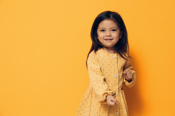 an adorable, playful little girl in a summer dress stands on an orange background with an empty space for an advertising mockup, making an emotional face