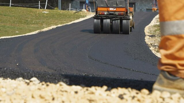 Asphalt roller compacting and levelling freshly laid asphalt surface on roadway. Operating construction machinery while finishing asphalting inclined driveway with bend in beautiful morning light.